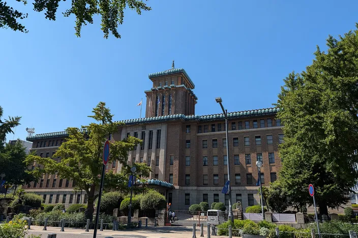 A long 1920s stone-and-brick public building with a squat tiered tower rising from its center, seen across a wide street from under trees on a cloudless summer day, with flagpoles and low hedges along its front.