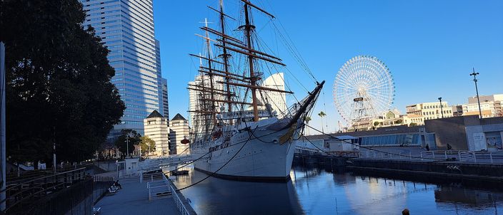 A white four-masted sailing ship moored in a stone dock in low winter sun, her bare masts and rigging against a clear blue sky, with a large white Ferris wheel and glass office towers standing behind her and the dock water still in the foreground.