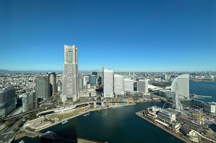 Elevated daytime view over Yokohama's Minato Mirai: the tapering Landmark Tower and a cluster of glass office towers along a harbour inlet, with the Cosmo Clock Ferris wheel on the right and blue sea and sky beyond