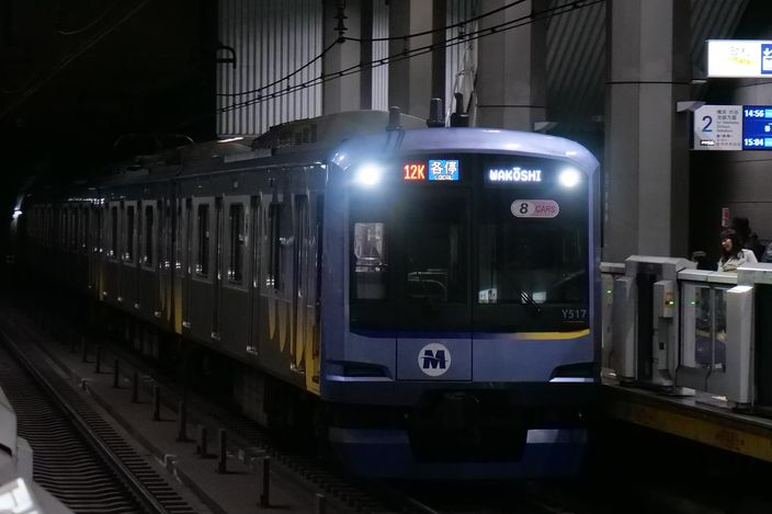 A blue Minatomirai Railway Y500 series train with an 'M' logo arriving at Shin-Takashima station platform, headlights on, in dim underground light