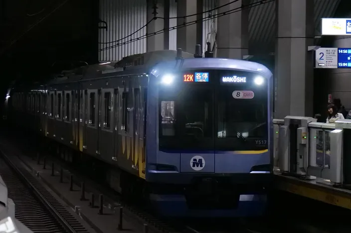 A blue Minatomirai Railway Y500 series train with an 'M' logo arriving at Shin-Takashima station platform, headlights on, in dim underground light