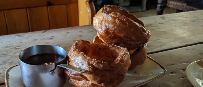 Two tall, crisp Yorkshire puddings on a pub plate beside a small jug of gravy on a wooden table