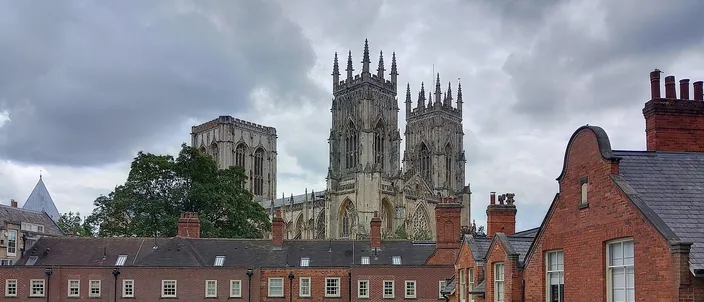 The west towers and central tower of York Minster rising behind the red-brick houses and chimneys of the Minster precinct under a gray, clouded sky, seen from the city walls
