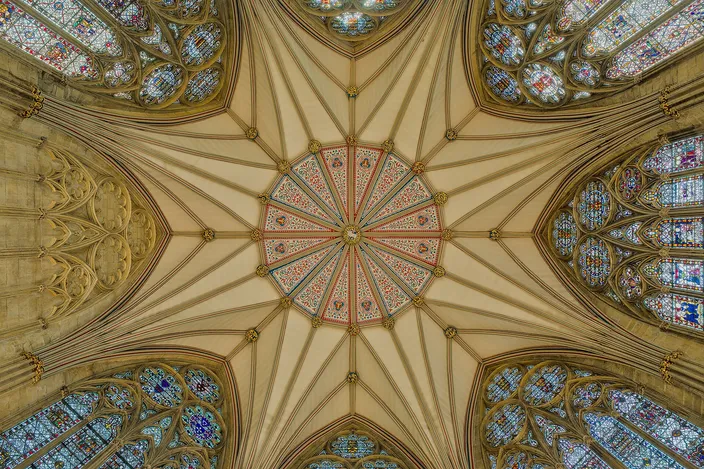 Looking straight up into the star-shaped vault of York Minster's octagonal Chapter House, ribs radiating from a painted central boss with tall stained-glass windows around the edge
