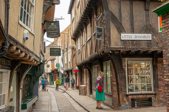 The Shambles in York, a narrow stone-paved lane of timber-framed houses with overhanging upper floors and hanging shop signs, a Little Shambles street sign on the right and a few walkers