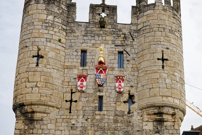 The upper stages of Micklegate Bar, York's southern gate, two round corner turrets in pale stone with painted coats of arms on the face between them