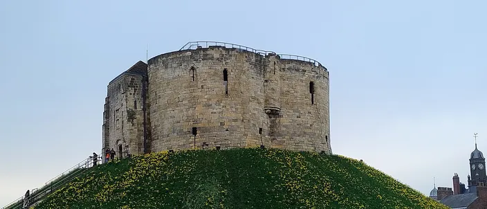 Clifford's Tower, the round stone keep of York Castle, on top of its steep green mound under a pale sky, with a church spire and rooftops at the edge