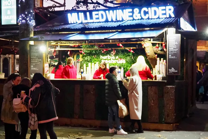 A wooden chalet at York's Christmas market at dusk under a lit sign reading Mulled wine and cider, staff serving behind the counter and visitors in front