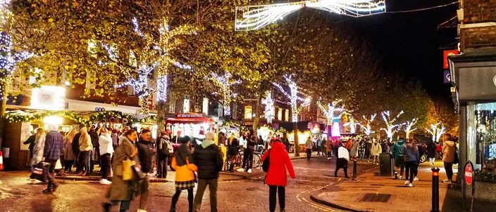 The Christmas market on Parliament Street in York at dusk, wooden chalets lit along a wide cobbled street under trees strung with lights, crowds walking between them