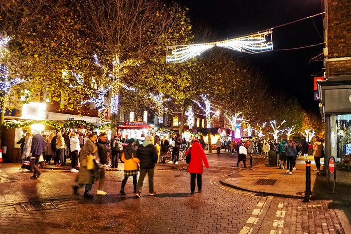 The Christmas market on Parliament Street in York at dusk, wooden chalets lit along a wide cobbled street under trees strung with lights, crowds walking between them