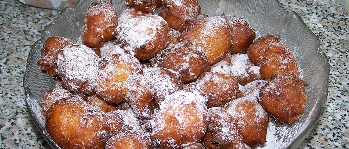 A glass dish heaped with small round fritule under icing sugar