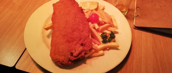A breaded veal parcel on a white plate with chips, a lemon wedge and a slice of pickle