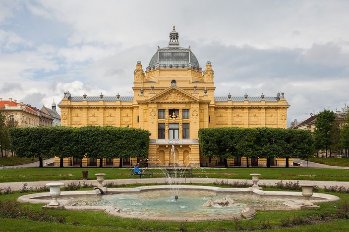 The Art Pavilion in Zagreb seen head-on across a lawn: a symmetrical mustard-yellow neo-classical hall with a columned centre and a dark dome, with a low circular stone fountain playing in front of it under a cloudy sky