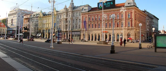 Trg bana Jelacica in low evening light: pastel Habsburg facades with ornate roofs along the far side of a wide paved square, a green digital clock mounted on a rooftop, tram wires overhead and empty tram tracks curving across the foreground