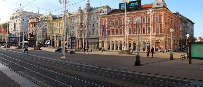 Trg bana Jelacica in low evening light: pastel Habsburg facades with ornate roofs along the far side of a wide paved square, a green digital clock mounted on a rooftop, tram wires overhead and empty tram tracks curving across the foreground