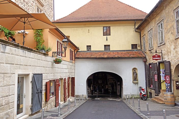 The Stone Gate in Zagreb's Upper Town seen from the cobbled lane below: a tunnel-like archway under a red-tiled roof between two old houses, a statue in a niche on the wall to the right and a cafe umbrella on the left