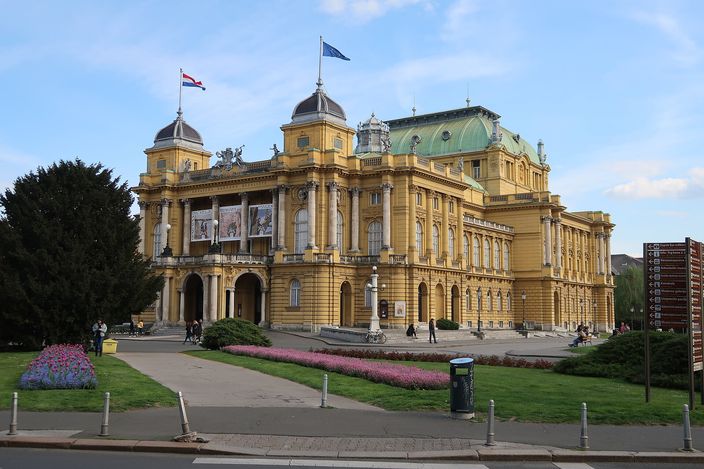 The Croatian National Theatre in Zagreb seen across a lawn: a symmetrical yellow neo-baroque opera house with two corner cupolas and a green central dome, flags flying from its roof and flowerbeds in front of it under a blue sky