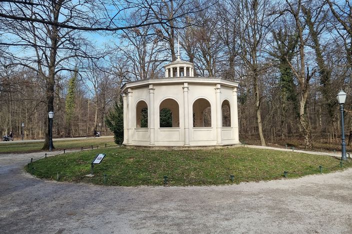 The Echo pavilion in Maksimir Park, Zagreb: a small round neo-classical pavilion of pale plaster with arched openings between columns and a low domed lantern, standing on a grassy mound among bare winter trees with gravel paths curving round it