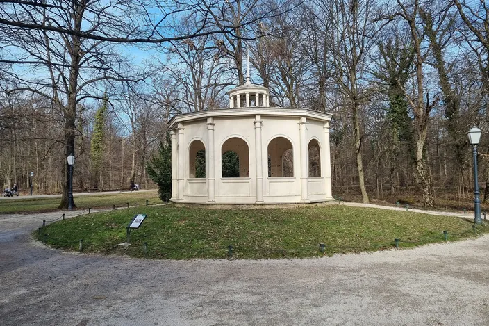 The Echo pavilion in Maksimir Park, Zagreb: a small round neo-classical pavilion of pale plaster with arched openings between columns and a low domed lantern, standing on a grassy mound among bare winter trees with gravel paths curving round it