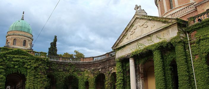 The arcades of Mirogoj cemetery in Zagreb: a curving colonnade of stone columns smothered in ivy, with a pedimented porch at the right beneath a large verdigris-copper dome and a smaller green dome further along the wall, under a grey clouded sky