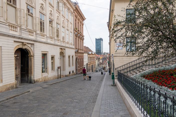 A quiet cobbled Upper Town street (Radićeva) lined with pale Habsburg façades, a person walking a dog, flower boxes along an iron railing, and a modern tower visible downhill