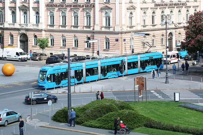 A bright blue low-floor Zagreb tram on a curve in the street, passing a long pale-pink 19th-century building, with cars, pedestrians and a clipped hedge in the foreground