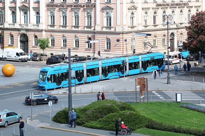 A bright blue low-floor Zagreb tram on a curve in the street, passing a long pale-pink 19th-century building, with cars, pedestrians and a clipped hedge in the foreground